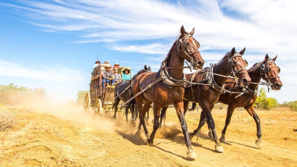 A horse-drawn stagecoach travels on a dusty trail, carrying passengers under a blue sky.