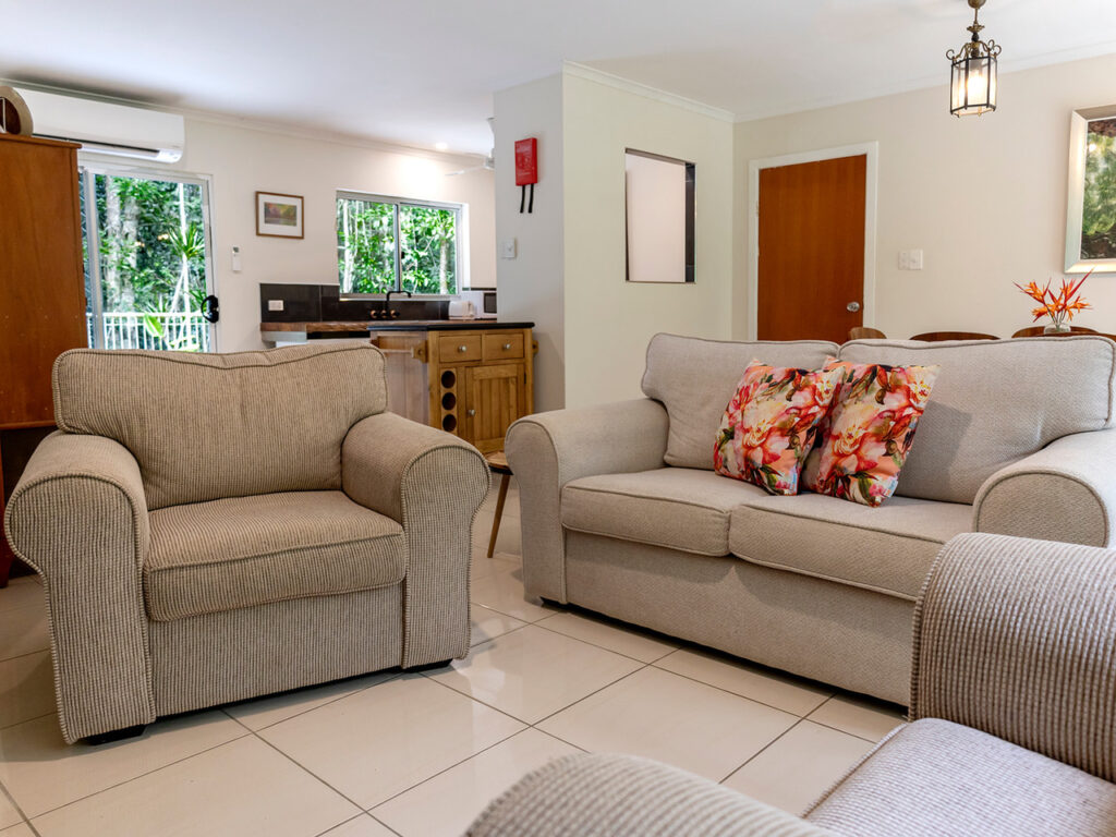 Cozy living room with beige armchair and sofa, decorated with floral pillows. Dining table and kitchen visible in the background.