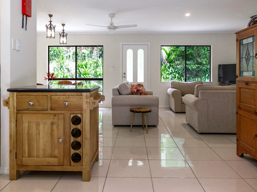 Open-plan living room with tiled floor, featuring two beige sofas, a small wooden table, and a TV. There is a kitchen island with wine storage on the left and large windows overlooking greenery.