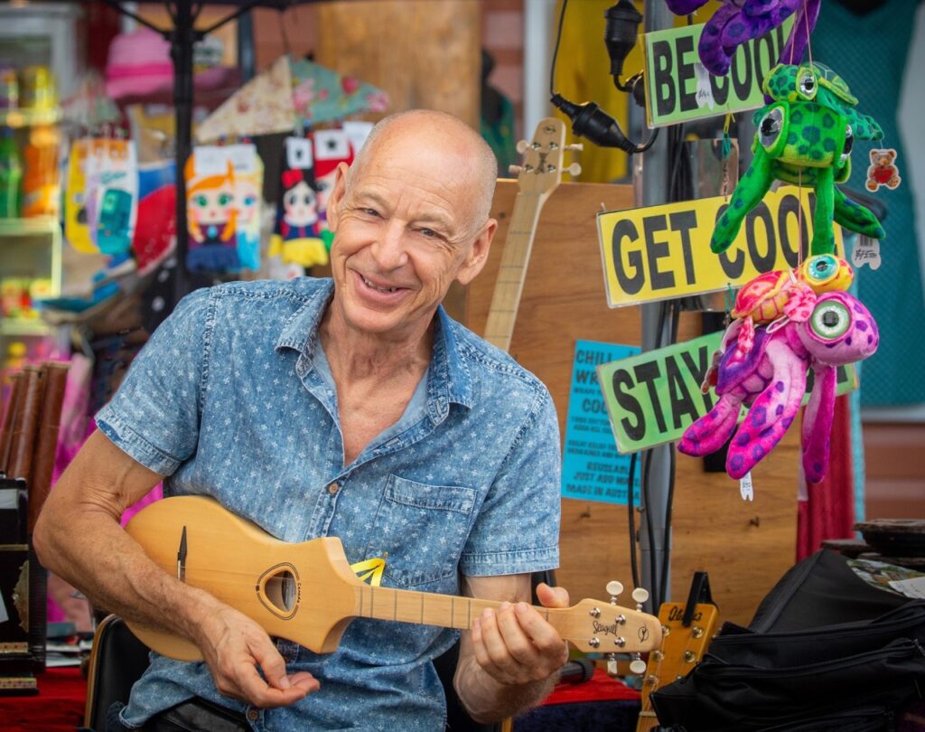 A man in a blue shirt plays a small guitar at a colorful market stall displaying plush toys and various signs.