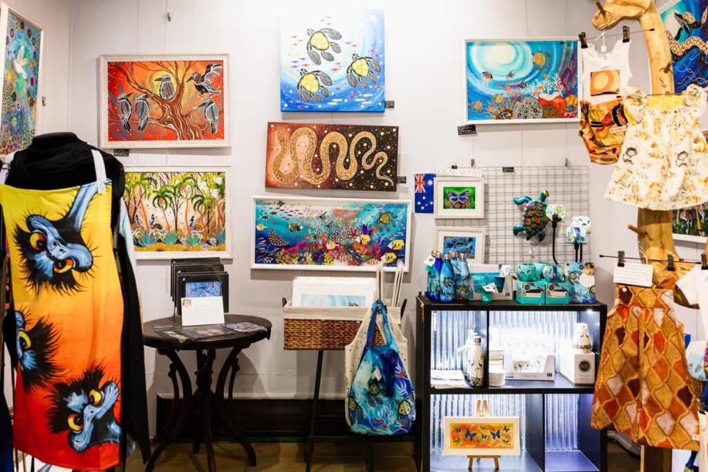 An art display featuring colorful paintings, textile designs, and various crafts in a well-lit room.
