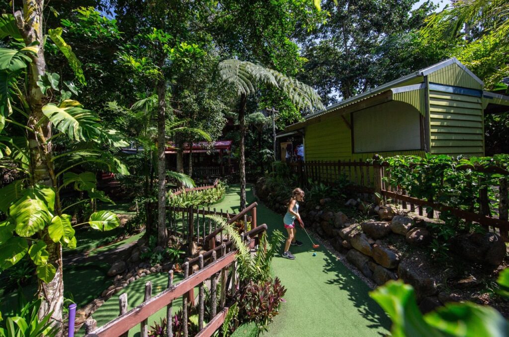 A person plays mini golf in a lush, tropical-themed course with plants and wooden structures surrounding the area.