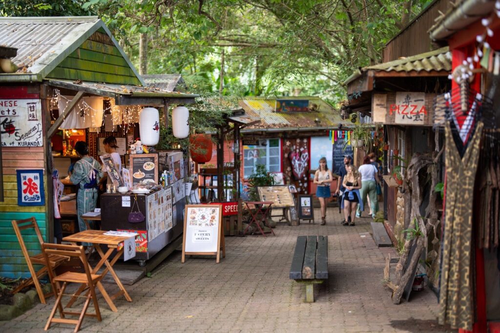 A quaint outdoor market with small shops and food stalls, surrounded by trees. Two people walk down the pathway in the background.