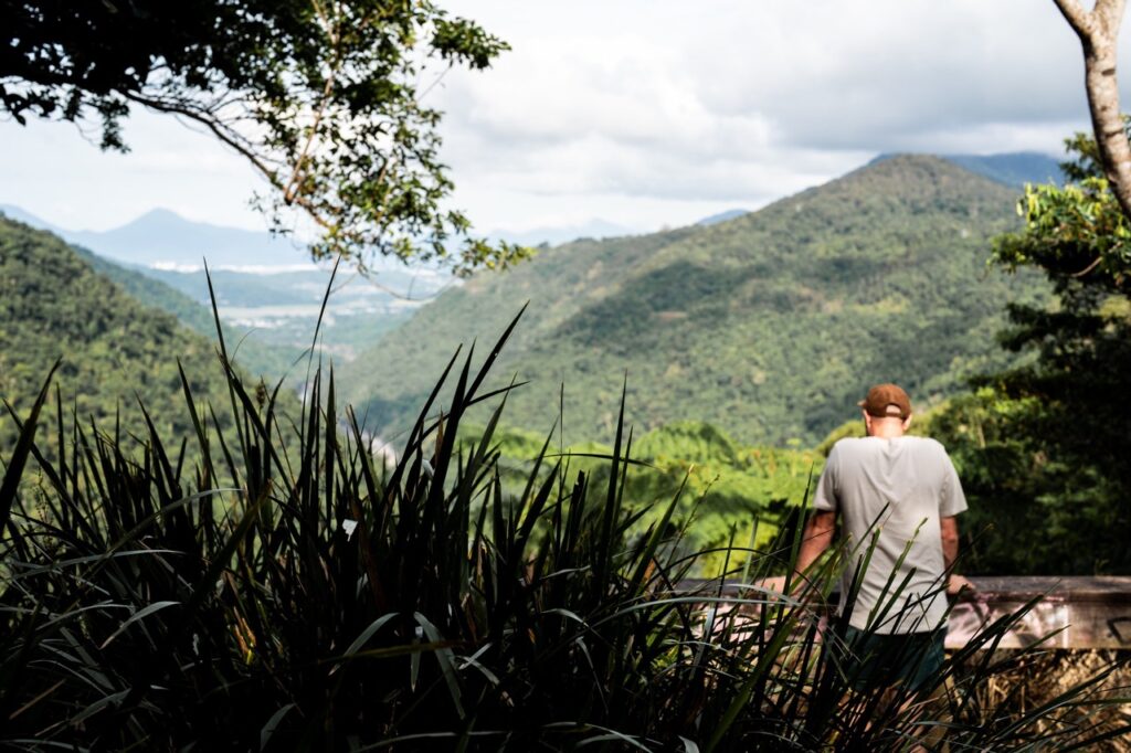 A person wearing a cap stands near a railing, overlooking a lush valley with green hills under a cloudy sky.