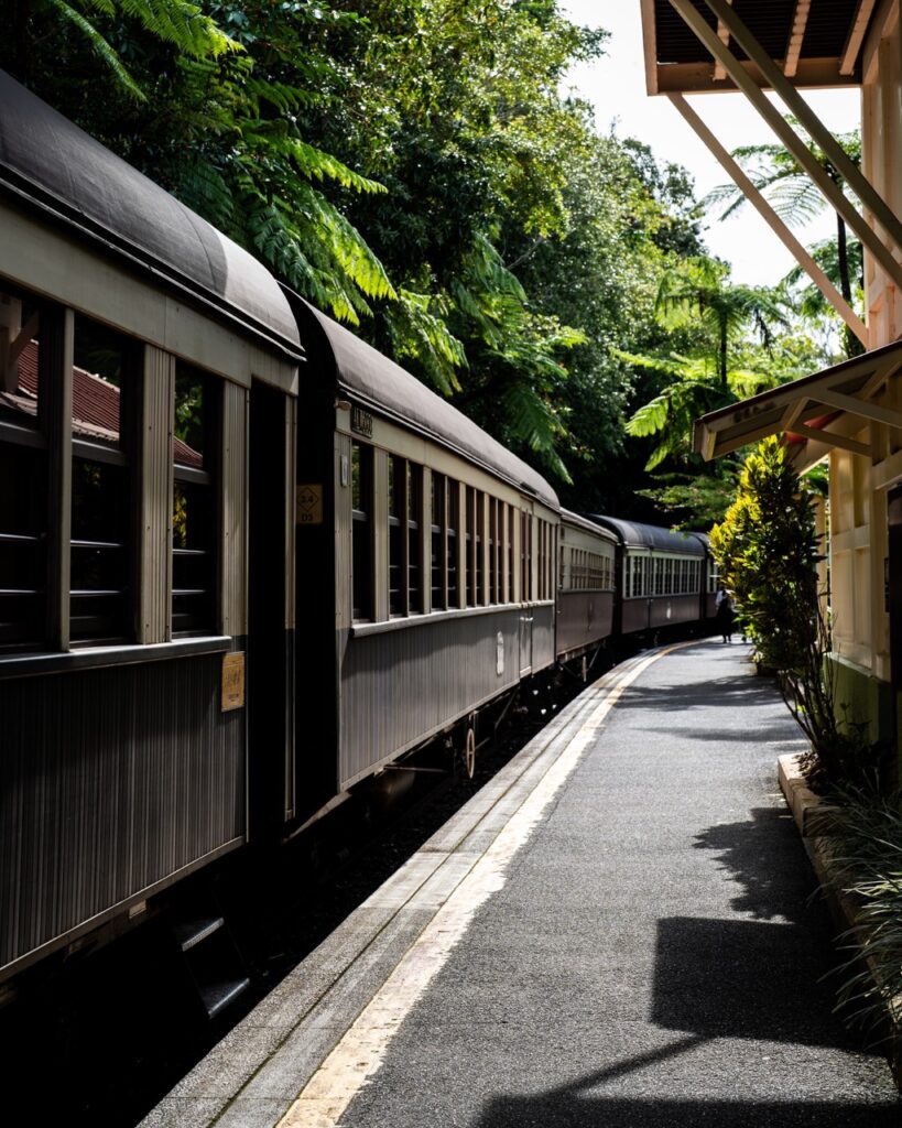 A vintage train moves along a curved, sunlit track next to a platform shaded by lush green trees.