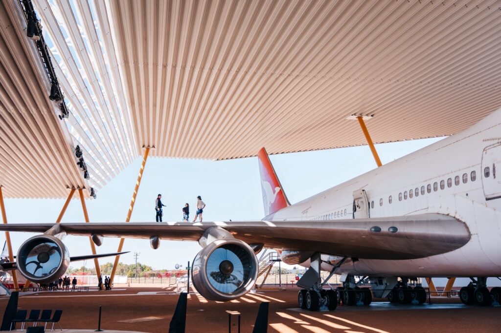 Two people stand on the wing of a large passenger airplane under a modern canopy structure with yellow supports.