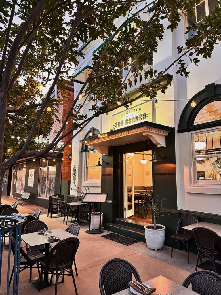 A charming outdoor dining area with tables and chairs is set up in front of a restaurant with warm lighting and a sign that says 