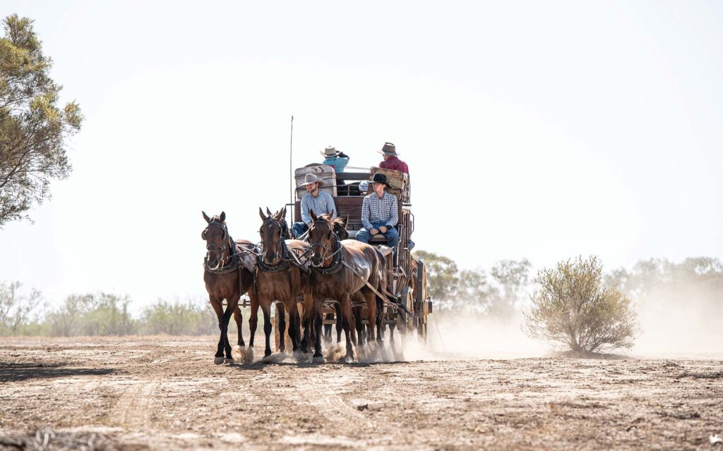 A horse-drawn wagon with people riding on it travels down a dusty road in a flat, arid landscape.