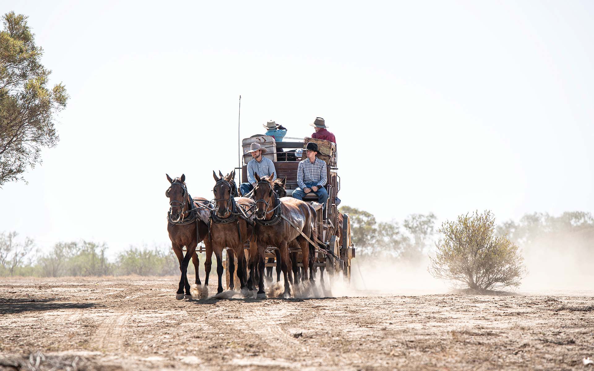 A horse-drawn wagon with people riding on it travels down a dusty road in a flat, arid landscape.