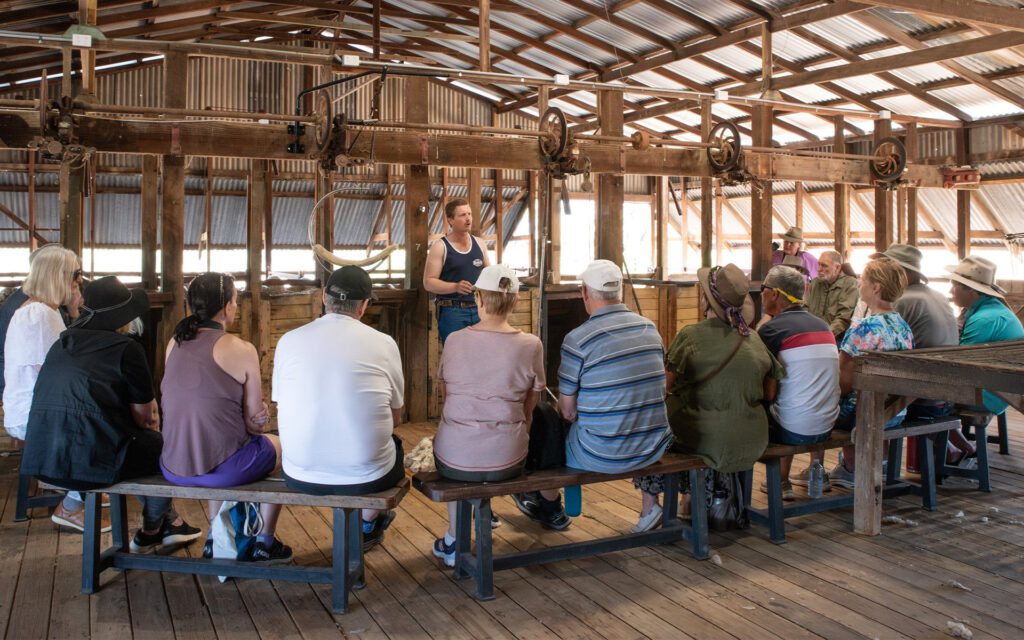 A group of people sit on benches listening to a person speaking in a rustic, wooden building with a high ceiling.