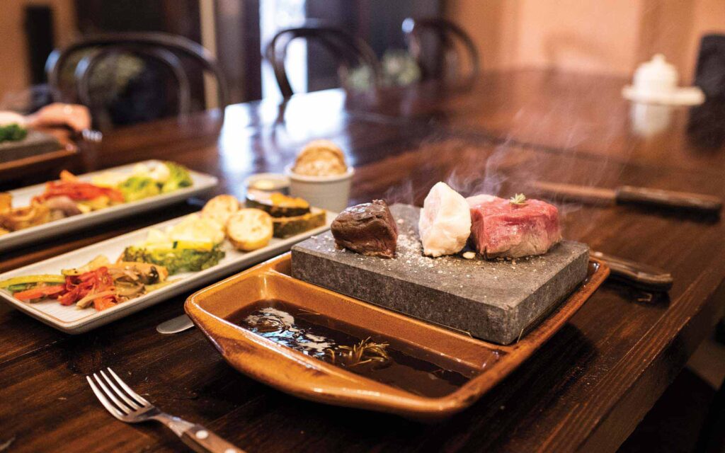 Steak and vegetables on a hot stone cooking plate with dipping sauce on a wooden table, accompanied by a side of grilled vegetables and bread.