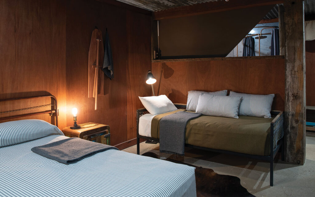 A cozy, dimly lit bedroom with two neatly made beds, wooden walls, a hanging robe, and a bedside lamp.