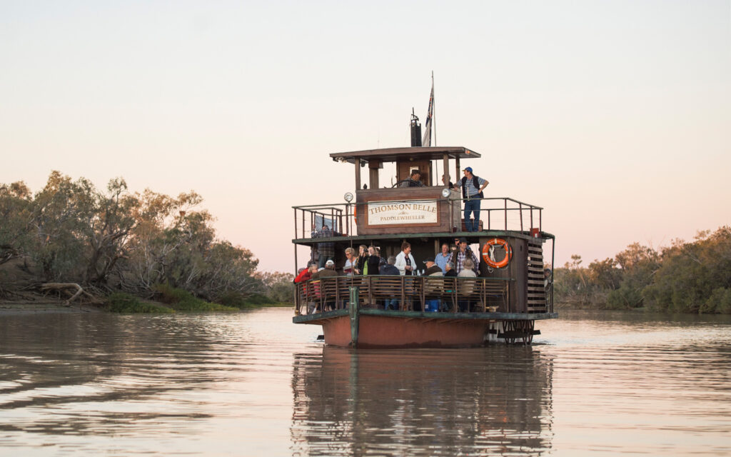 A riverboat with passengers cruises on calm waters surrounded by trees under a clear sky.