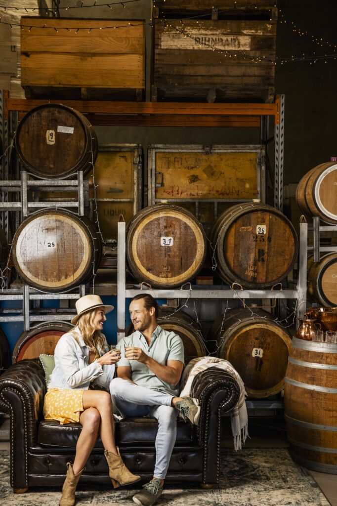 A man and a woman sit on a leather couch clinking glasses in a room filled with wooden barrels.