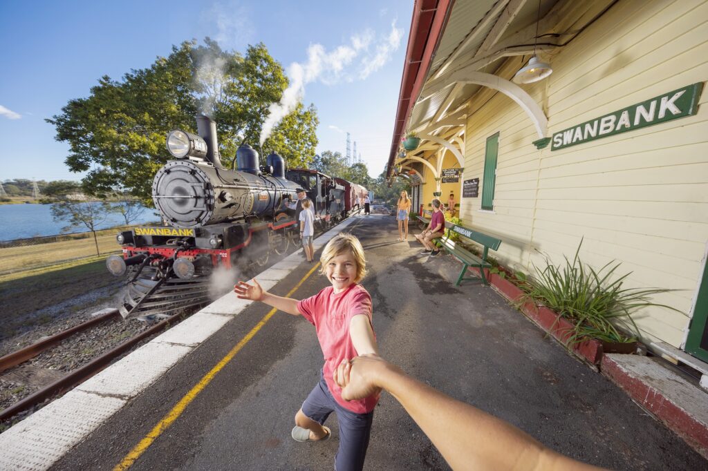 Child joyfully leading the viewer by hand at Swanbank train station as a vintage steam train arrives, with onlookers on the platform.