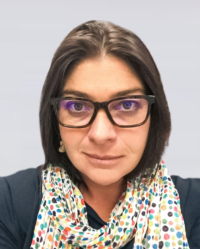 A woman with glasses and dark shoulder-length hair wearing a polka dot scarf and dark clothing against a plain background.