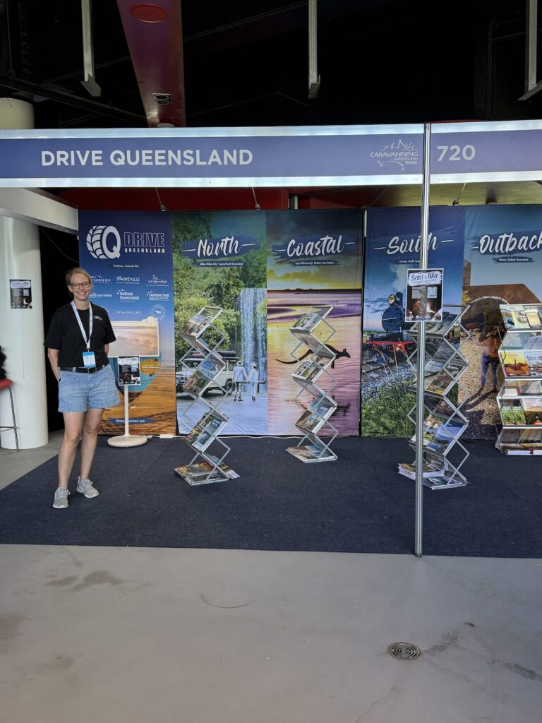 A woman stands at the Drive Queensland booth, featuring brochures and large scenic posters labeled North, Coastal, South, and Outback, at an indoor expo.