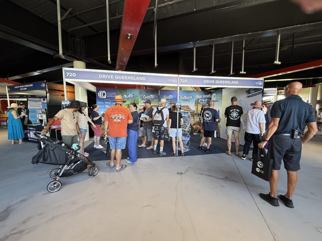 People stand and gather at the Drive Queensland booth inside a large indoor venue; a stroller and various signage are visible.