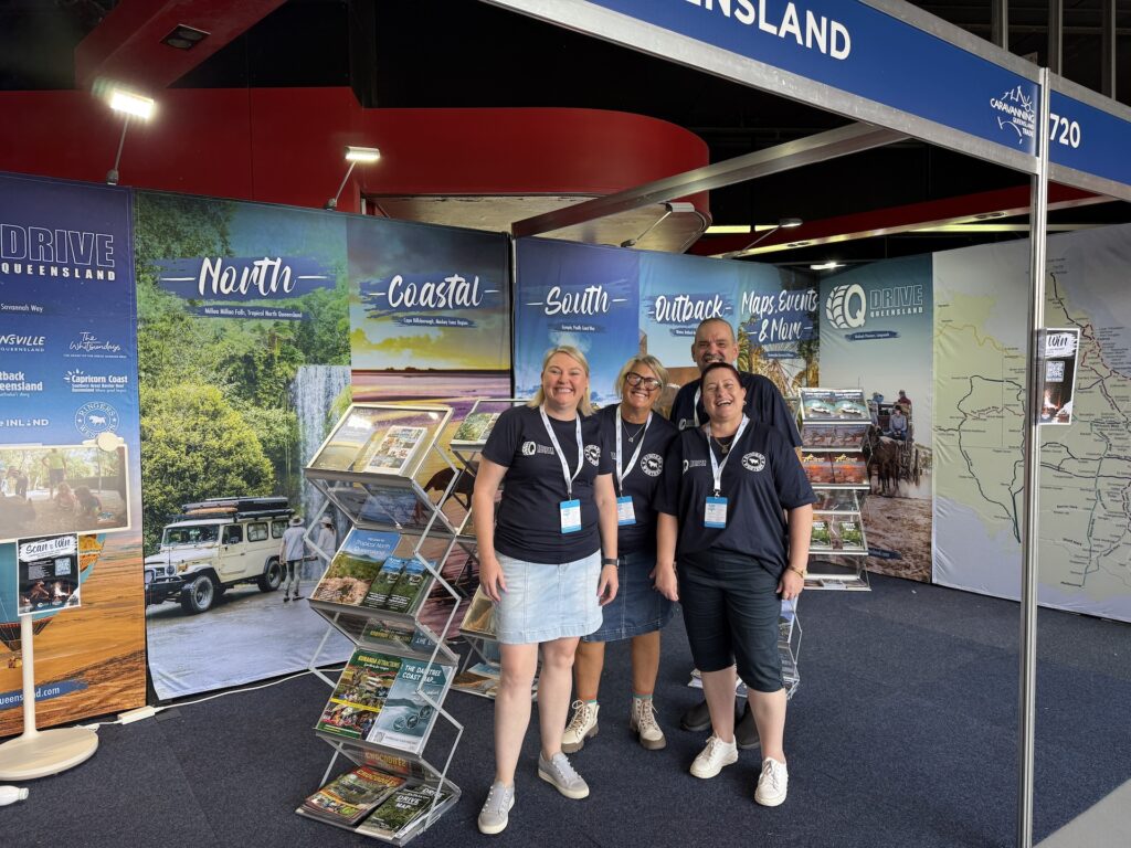 Four people wearing matching navy shirts stand and smile at a tourism booth featuring maps, brochures, and scenic Queensland travel displays.