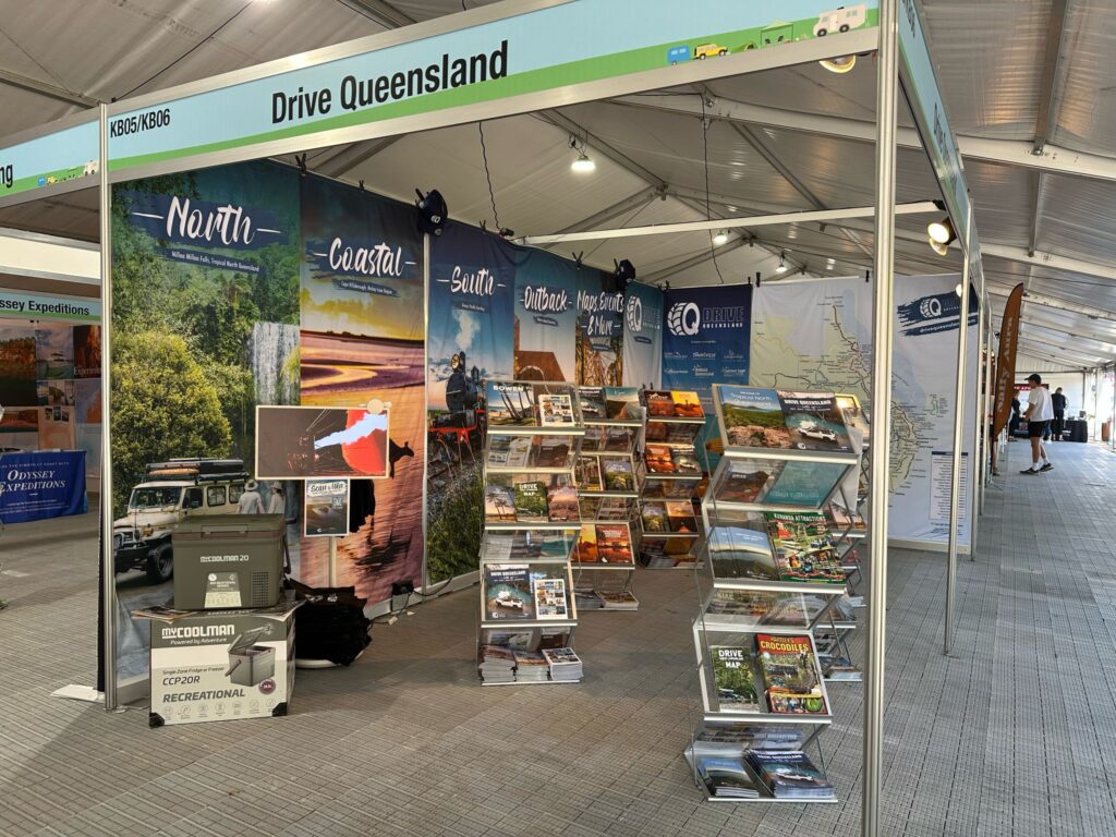 Drive Queensland exhibition booth with large scenic banners, brochure racks displaying travel guides, and a portable fridge, set up inside a large event tent.