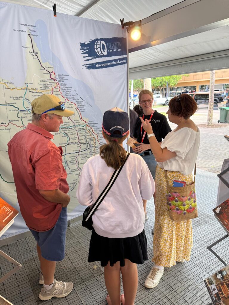 Four people stand in front of a large Queensland map display at an indoor event, engaged in conversation. One person appears to be explaining or pointing something out.