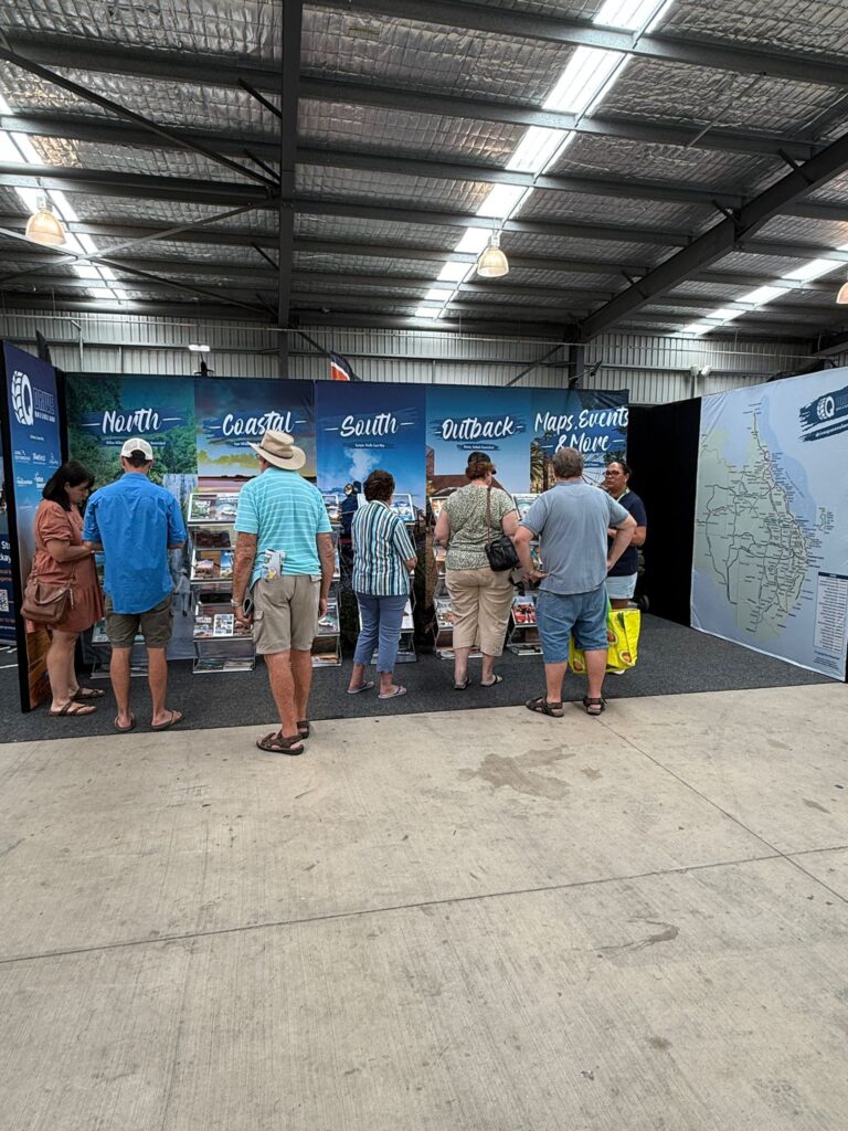 A group of people stands in front of a tourism information booth with regional maps and brochures inside an exhibition hall.