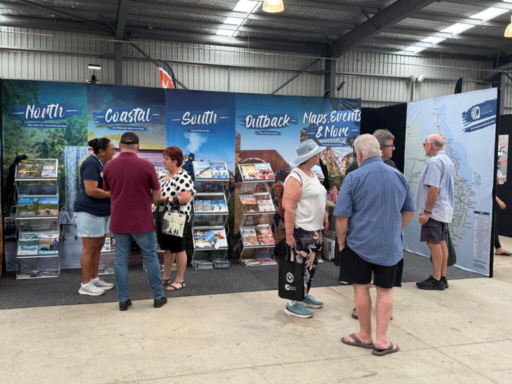 People browse and discuss brochures at a travel information booth divided into sections: North, Coastal, South, Outback, and Maps, inside a large indoor venue.