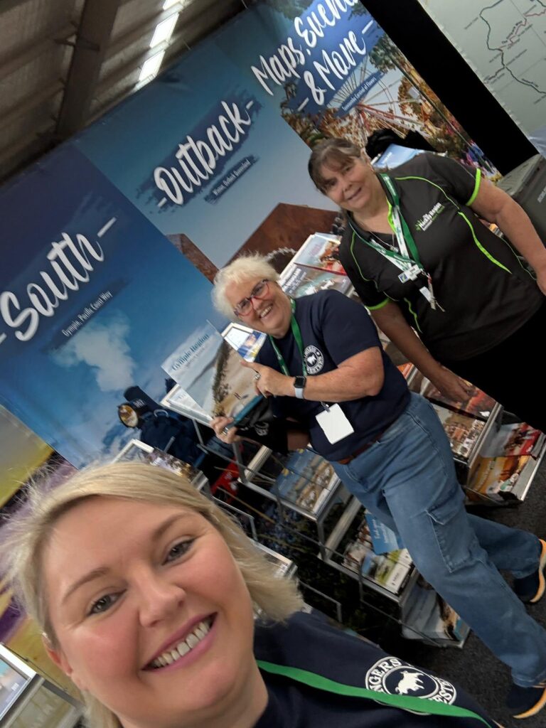 Three women stand smiling at a tourism booth featuring maps and brochures, with banners displaying 