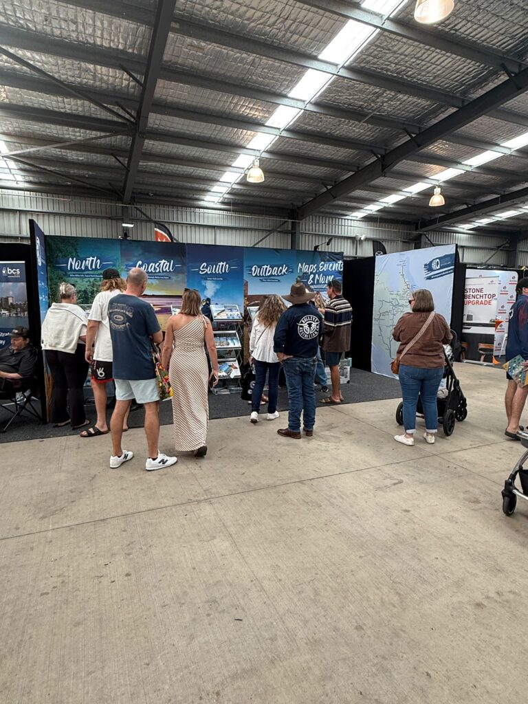 People standing and walking at an indoor expo or market, visiting booths with large banners and displays under a metal roof.