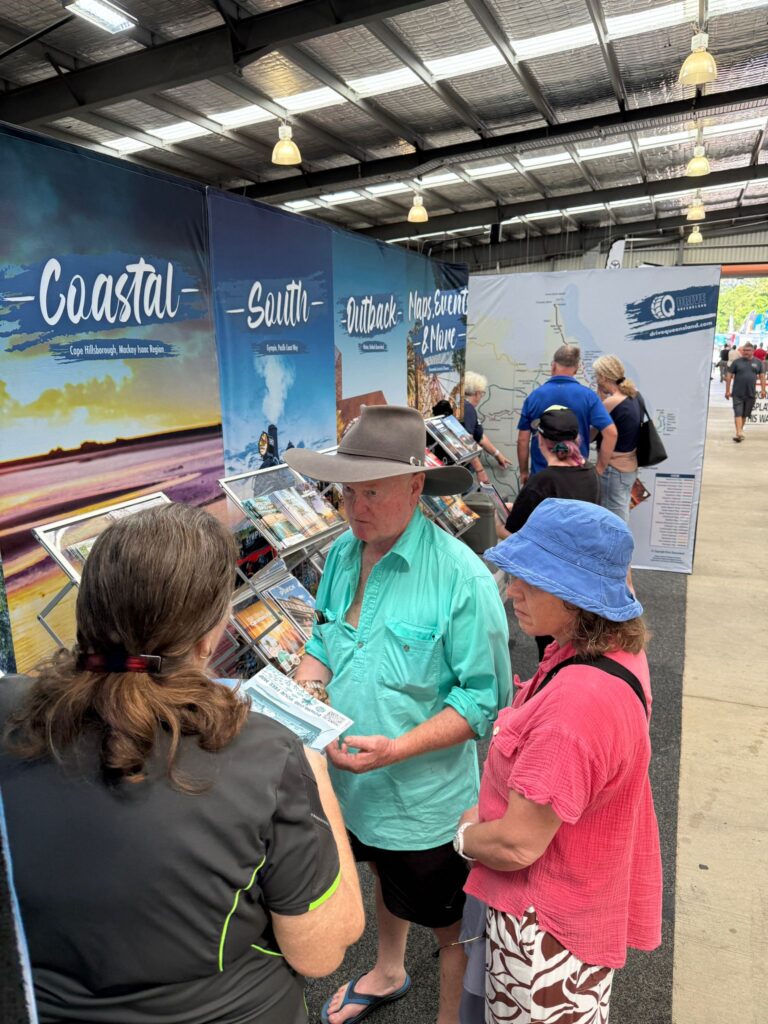 A man and a woman speak with an event staff member at a tourism booth inside an exhibition hall, with brochures and coastal displays in the background.