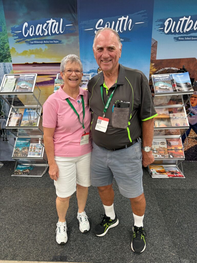 An older woman and man stand smiling together at an indoor event, wearing name badges and casual clothes, with travel brochures displayed behind them.