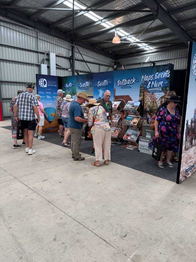 A group of people browse brochures and maps at a tourism information booth in an indoor venue with a high metal ceiling.