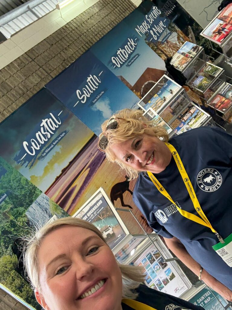Two women smiling at a tourism booth with banners labeled 