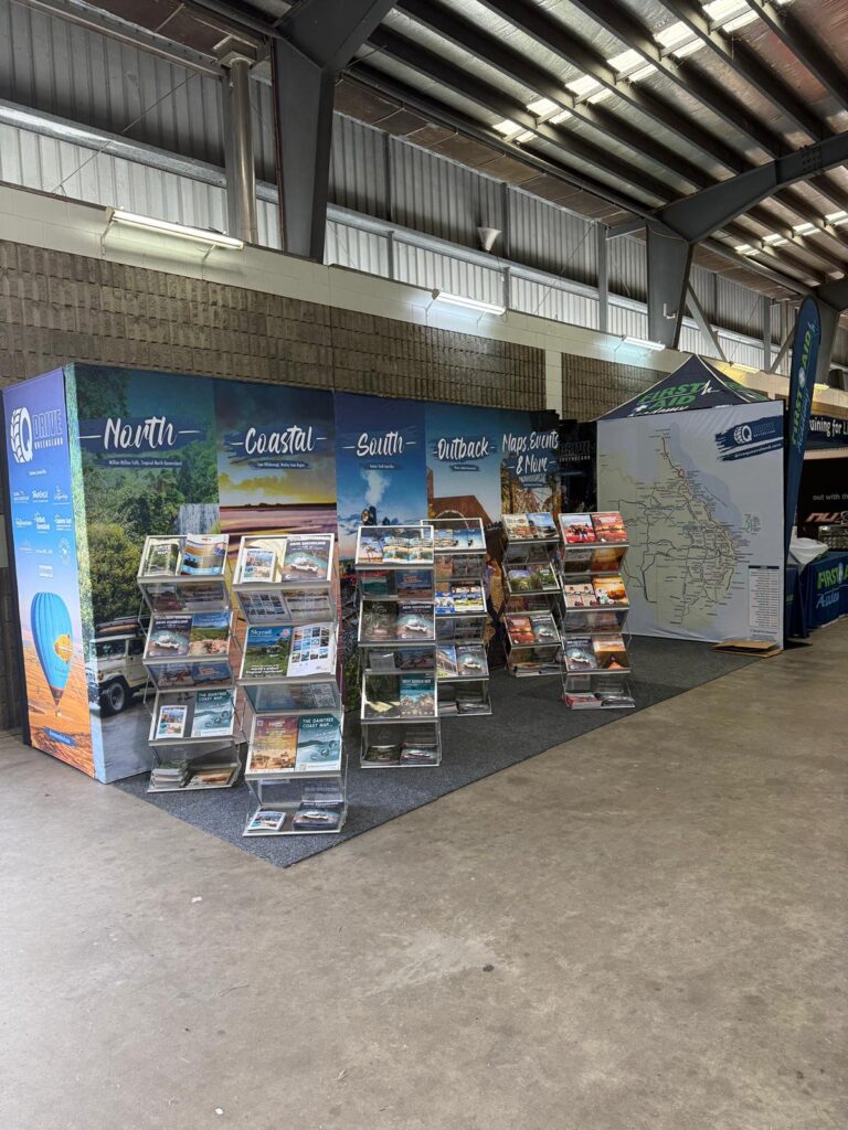 Indoor exhibition booth displaying travel brochures and maps on racks, with regional signage for North, Coastal, South, Outback areas, and a large map on the right wall.