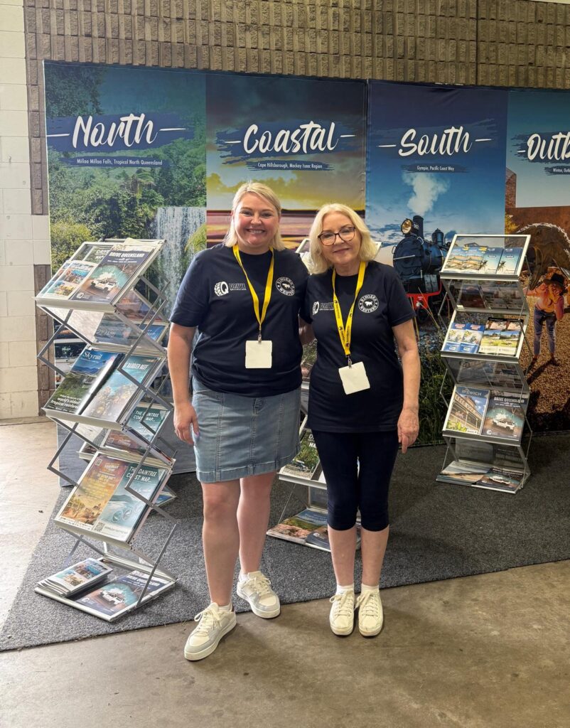 Two women wearing navy shirts and yellow lanyards stand in front of travel brochure displays labeled North, Coastal, South, and Outback.