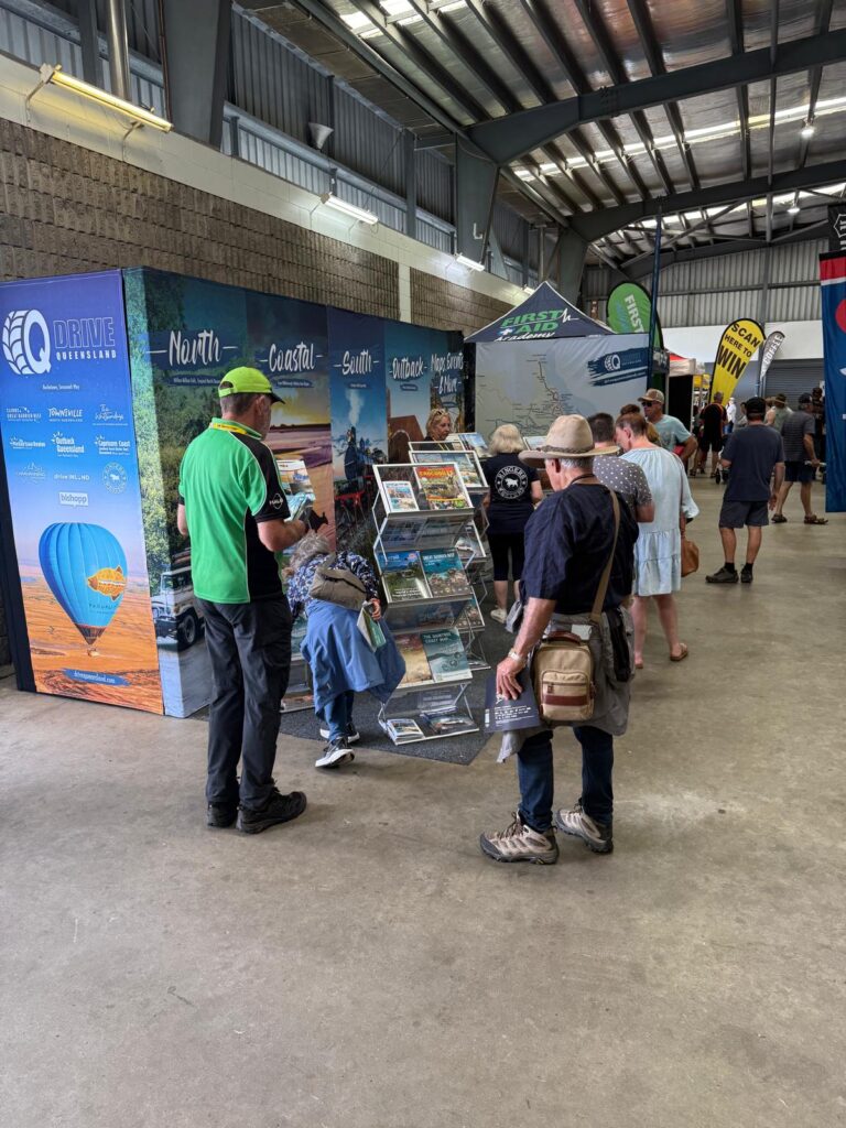 People browse pamphlets at a tourism information booth inside a large indoor venue with banners and displays in the background.