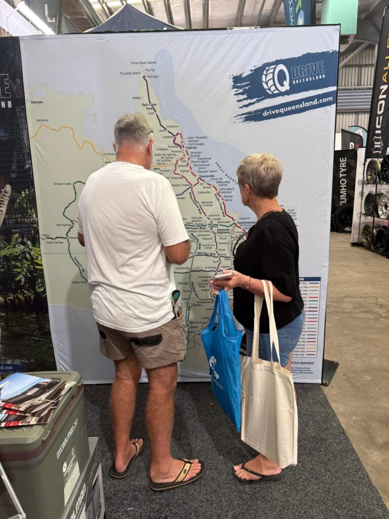 Two older adults stand in front of a large Drive Queensland map display, examining routes and holding shopping bags at an indoor event.