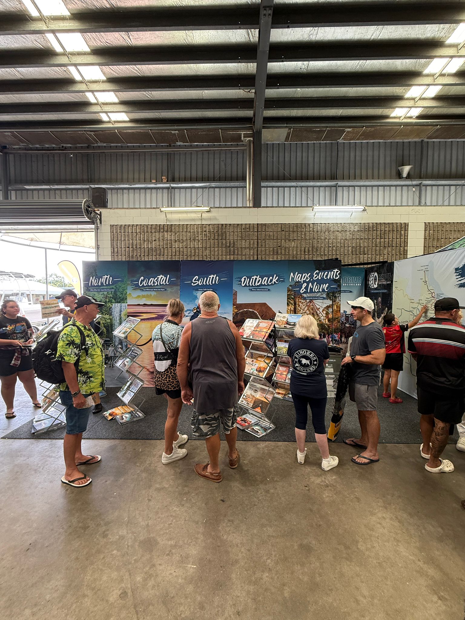 A group of people stand indoors at a display booth featuring maps and brochures organized by region, under a corrugated metal roof.