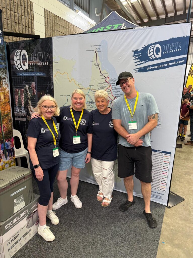 Four people stand in front of a Drive Queensland map display at an indoor event, wearing navy shirts and name lanyards, smiling at the camera.