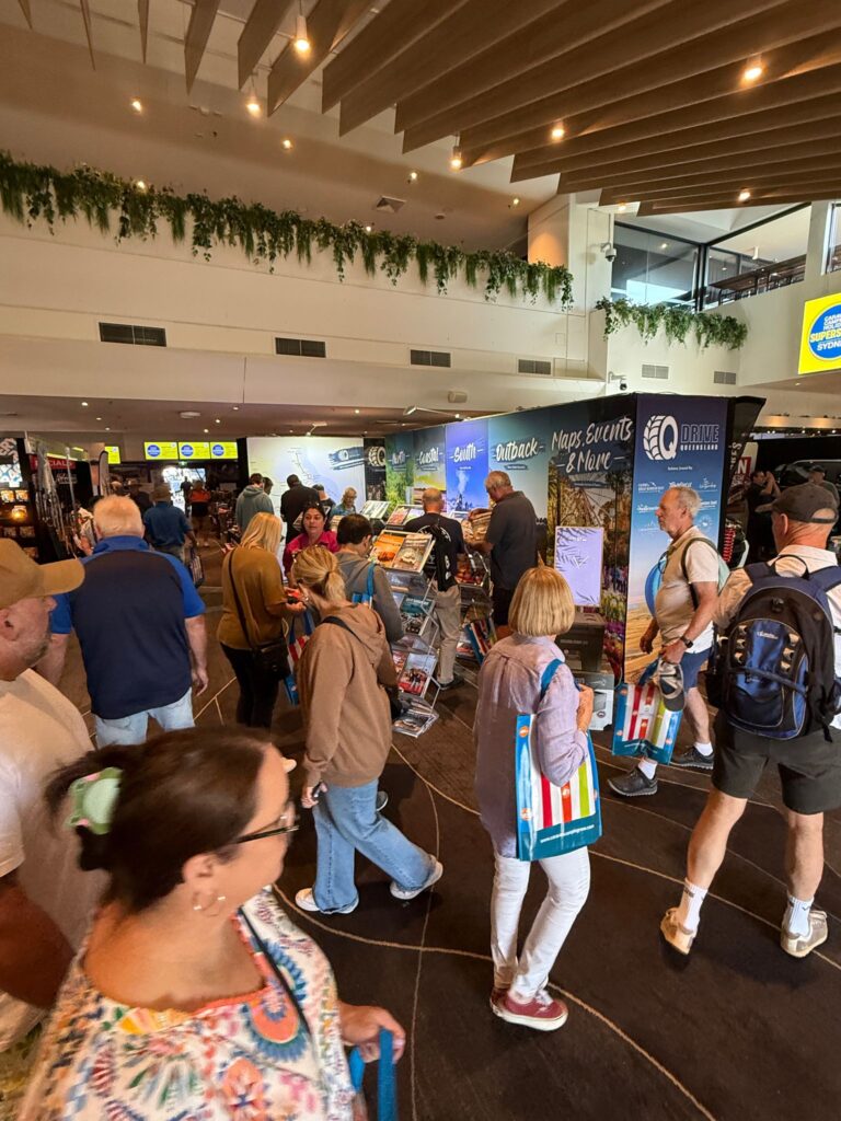 People walk through a busy indoor event or expo, browsing pamphlets and displays. Some attendees carry colorful bags. The atmosphere is lively and well-lit.
