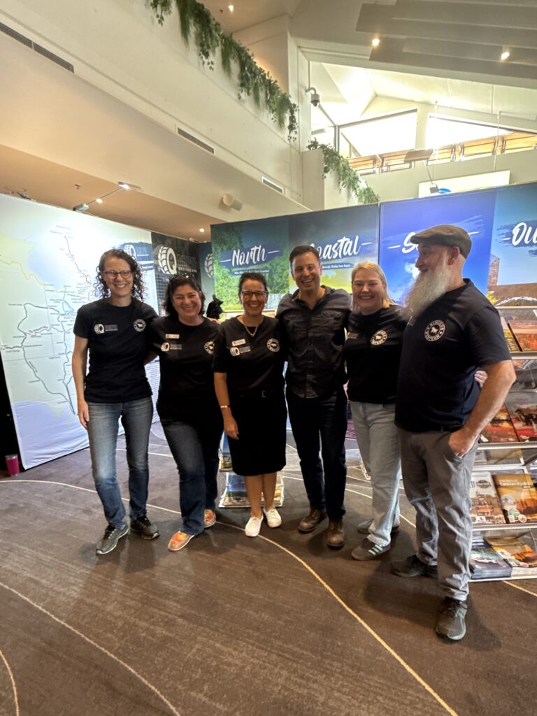 Six people stand together indoors, smiling at the camera in front of display boards with regional names and maps. They are wearing name tags and matching dark shirts.