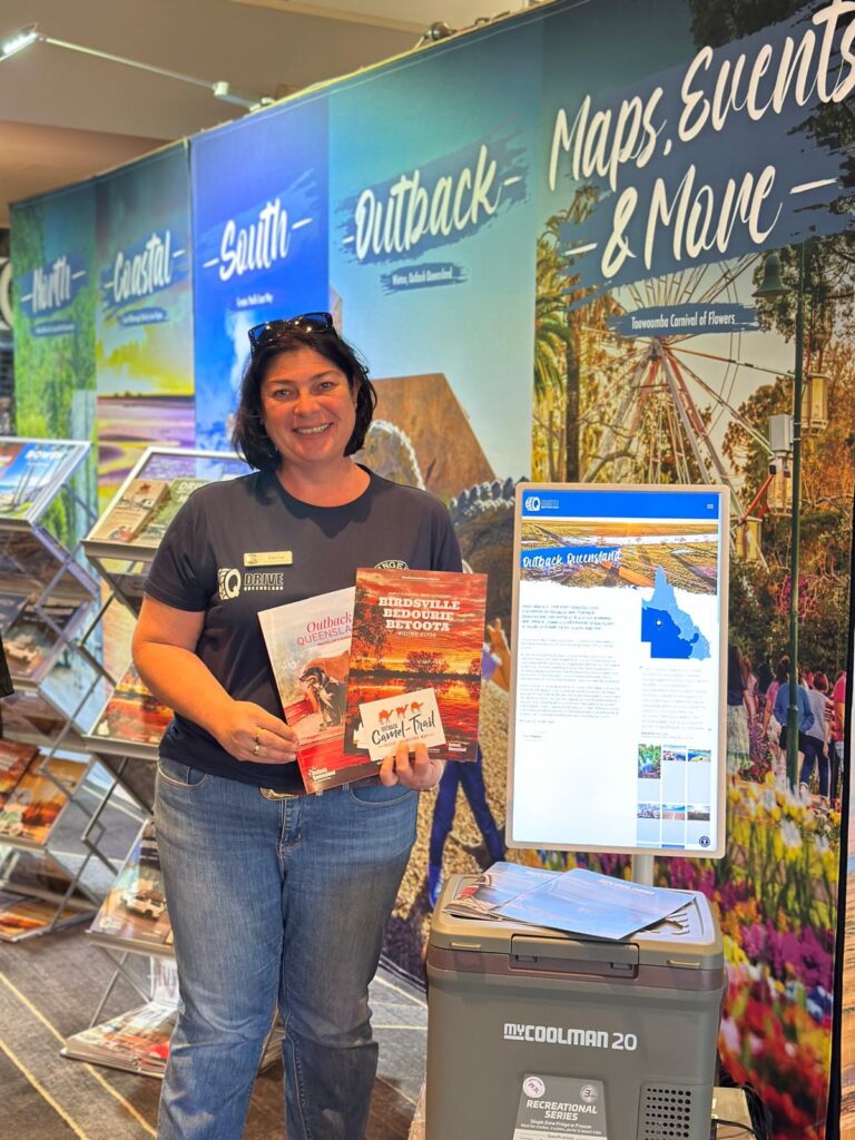 A woman stands indoors holding regional travel brochures next to a display with maps, event information, and tourism materials.