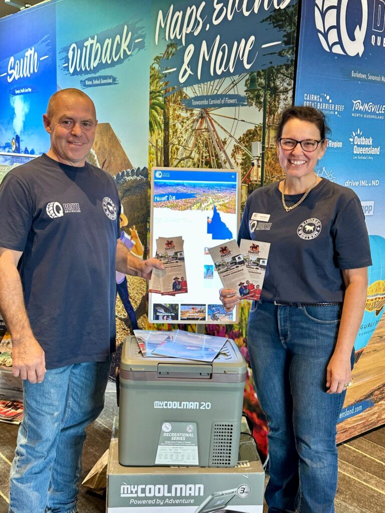 Two people wearing matching shirts stand indoors holding brochures in front of colorful travel displays and a portable cooler labeled 