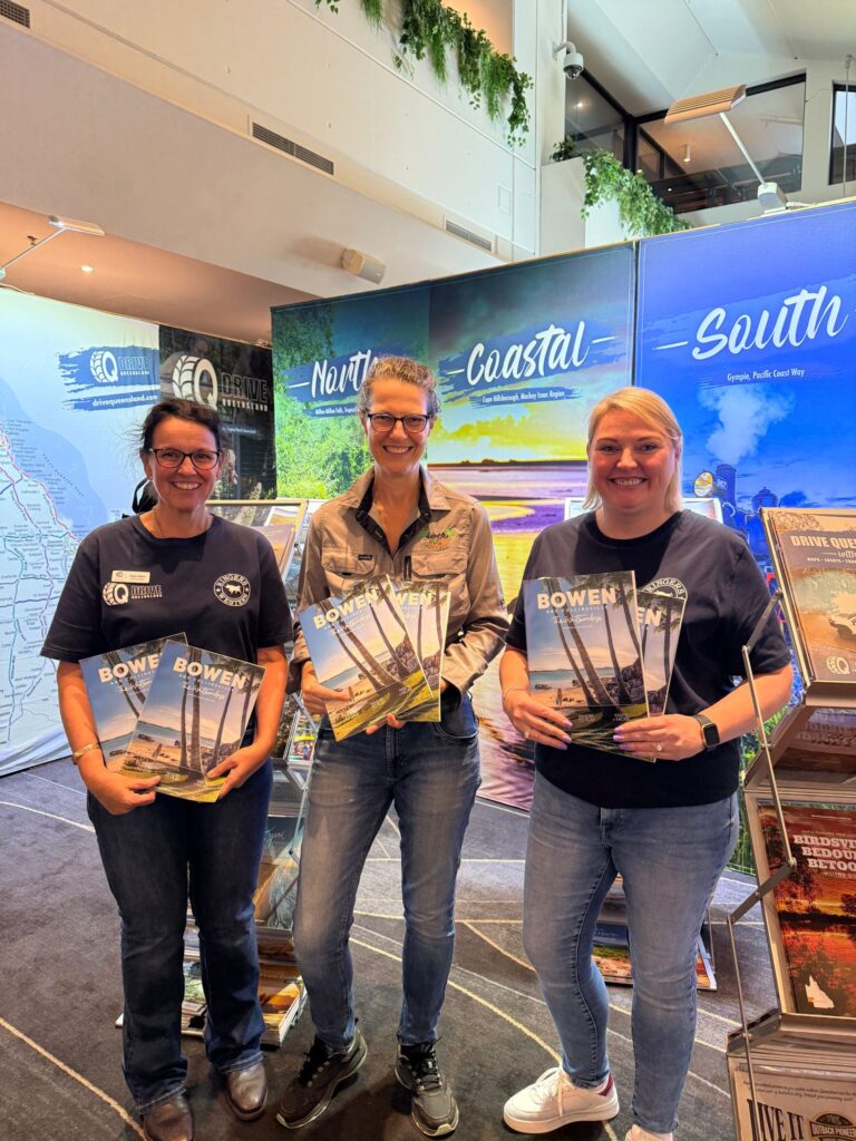 Three women stand indoors holding stacks of 