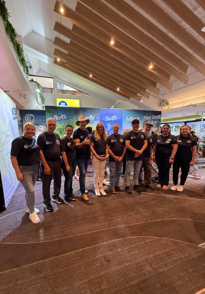 A group of eleven people in matching black shirts stands indoors on a carpeted floor, posing together in front of promotional banners.