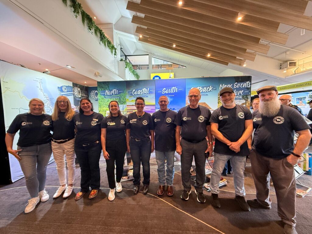A group of ten people wearing matching navy blue shirts stand in a row indoors, smiling at the camera. Informational banners and brochures are displayed behind them.