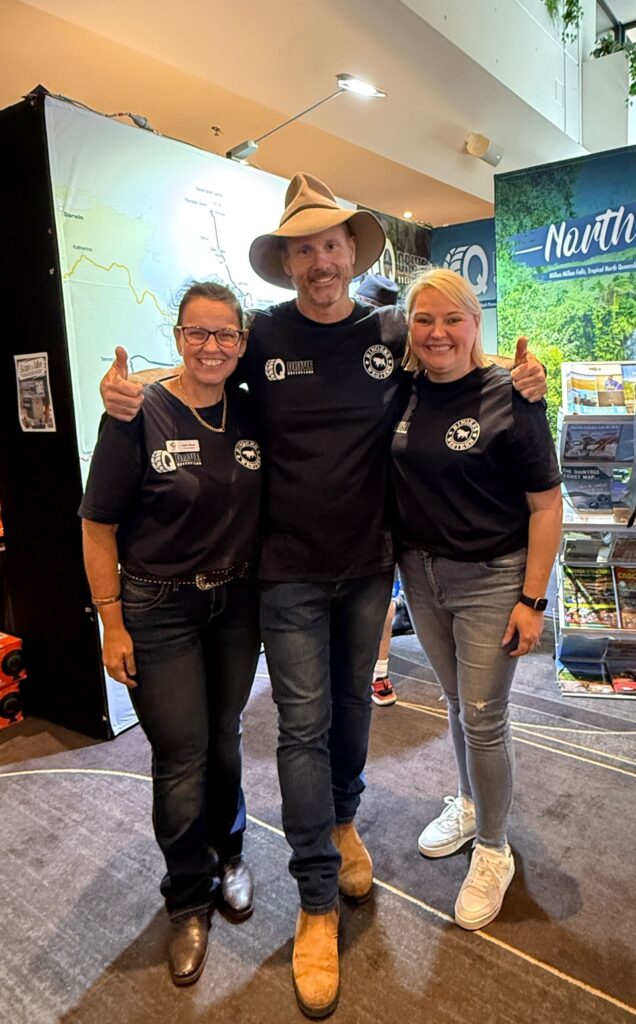 Three people stand together indoors, smiling at the camera. They are wearing matching black shirts with logos and casual jeans. A booth and brochures are visible in the background.