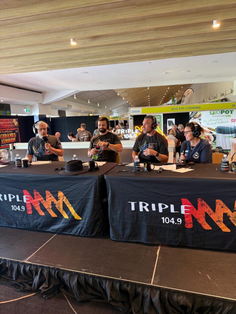 Four men sit at a table with Triple M 104.9 banners, wearing headsets and speaking into microphones, hosting a live radio broadcast in a public indoor space.