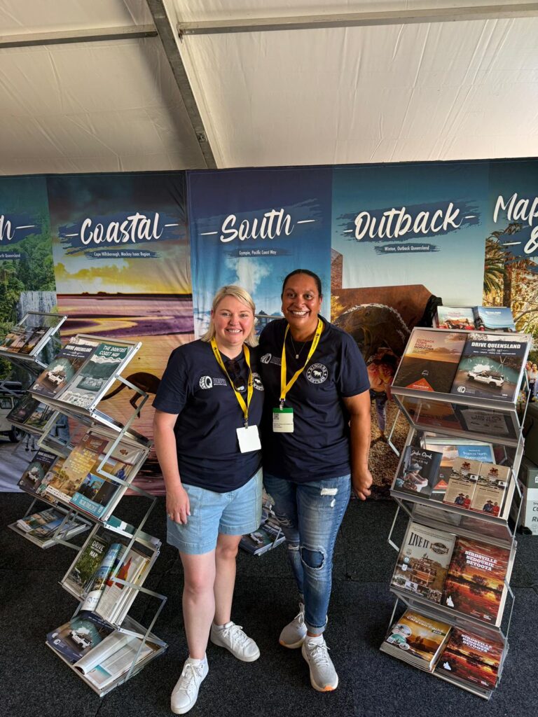 Two women stand smiling in front of travel brochure racks and large banners labeled 