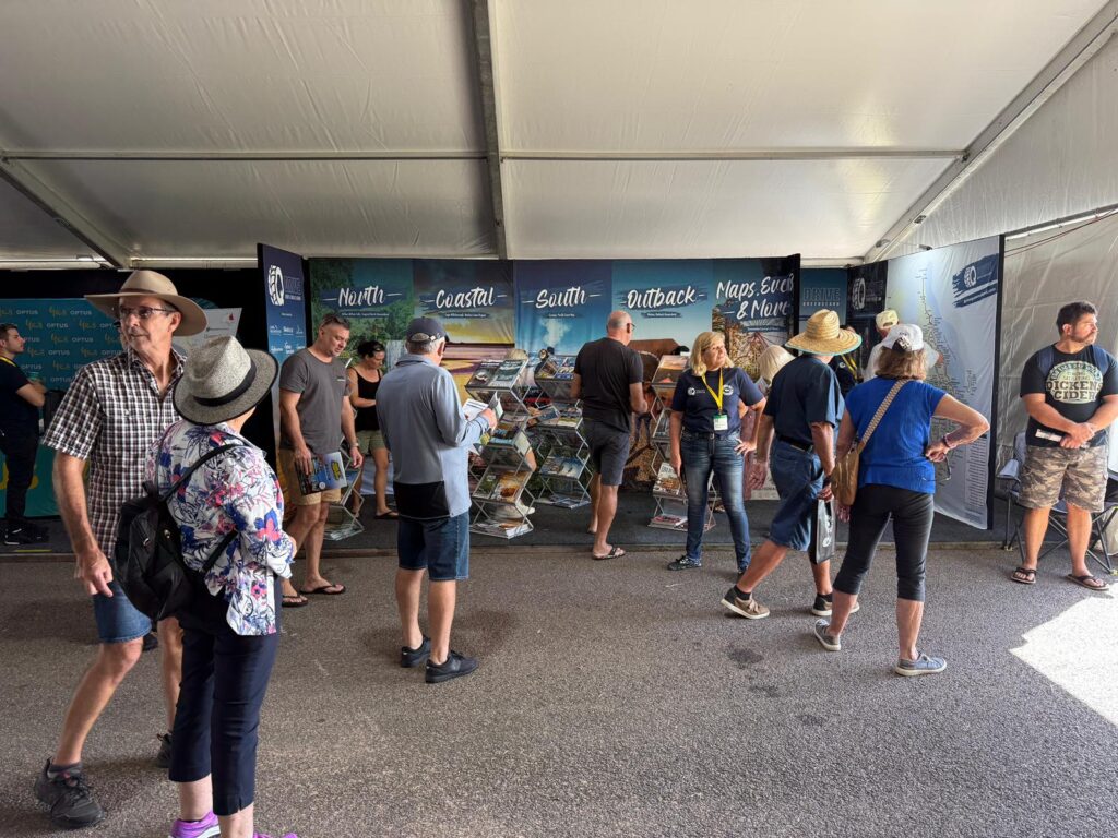 People stand and browse brochures at an indoor event booth with large regional signs on the back wall under a white tent.